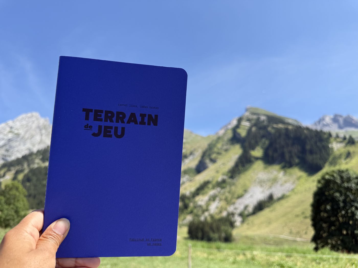 Carnet Terrain de Jeu bleu tenu à la main devant les montagnes, mention « Fabriqué en France, 46 pages »